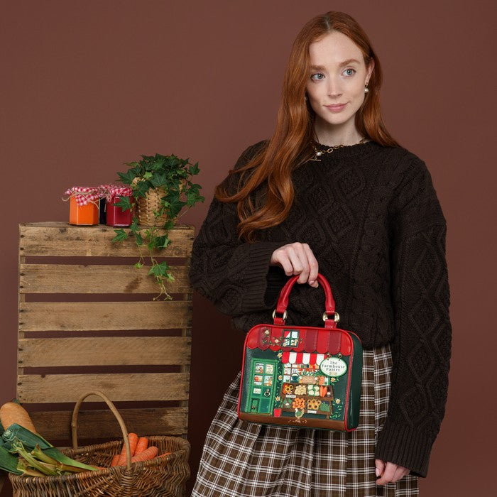 A woman with long red hair stands against a brown studio backdrop holding a small red handbag decorated with a colorful illustrated storefront scene. She wears a dark cable-knit sweater and a brown-and-white plaid skirt.