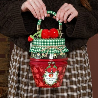A woman with long red hair stands against a brown studio backdrop holding a small decorative basket bag. The bag is red with green gingham trim, red strawberries, and a snowman face on the front.