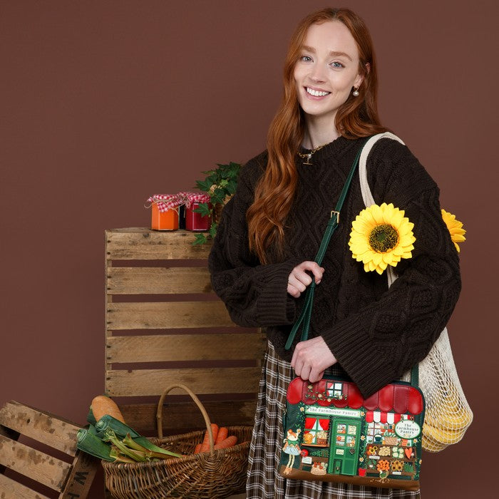 A smiling young woman with long red hair stands against a brown studio backdrop, wearing a dark cable-knit sweater and holding a decorative shoulder bag. The bag features a colorful Christmas village scene with red roofs, green doors, and a small white sign.