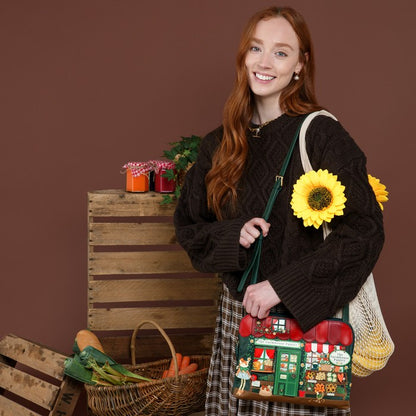 A smiling young woman with long red hair stands against a brown studio backdrop, wearing a dark cable-knit sweater and holding a decorative shoulder bag. The bag features a colorful Christmas village scene with red roofs, green doors, and a small white sign.