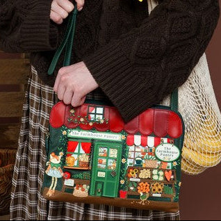 A smiling young woman with long red hair stands against a brown studio backdrop, wearing a dark cable-knit sweater and holding a decorative shoulder bag. The bag features a colorful Christmas village scene with red roofs, green doors, and a small white sign.