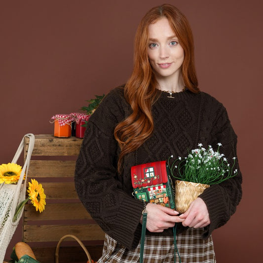 A woman with long red hair wearing a dark brown cable-knit sweater and a plaid skirt, holding a small red Christmas house purse and a potted baby's breath plant against a solid brown background.