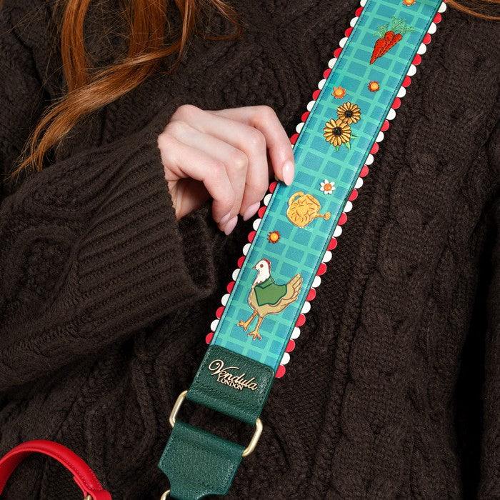 A close-up of a person wearing a dark brown cable-knit sweater, holding a decorative shoulder strap. The strap is teal with a plaid pattern and whimsical illustrations of a chicken, carrot, sunflower, and basket, finished with a dark green leather end and gold-tone hardware.