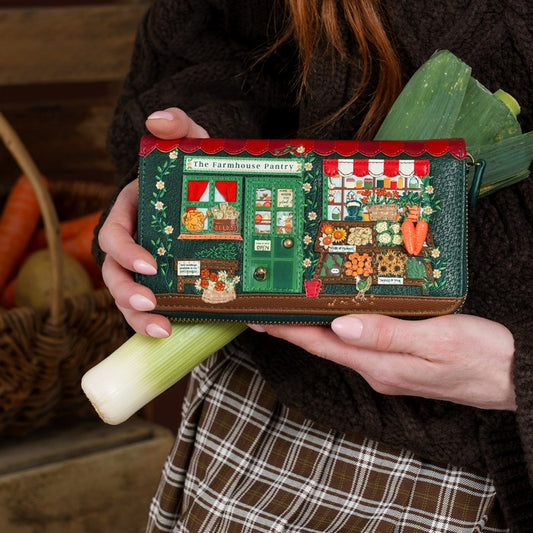 A person in a dark brown knit sweater holds a rectangular clutch wallet decorated with a whimsical illustrated farm market scene. The clutch features green foliage, red awnings, and colorful market stalls, with leeks and a basket visible in the softly blurred background.
