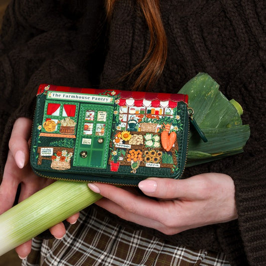 A person holds a small zip-around clutch shaped like a vintage bakery storefront with embroidered-style details, set against a dark brown knit sweater and green vegetable props.