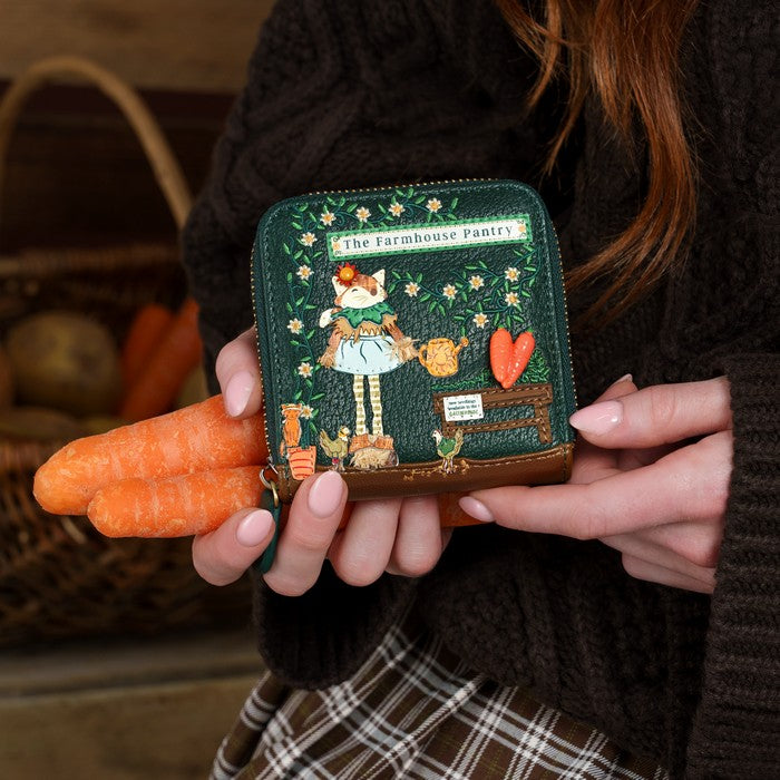 A person in a dark brown knit sweater holds a small embroidered wallet featuring a countryside pantry illustration. The wallet is dark green with floral embroidery, a cartoon girl, and a brown leather base, while the person also holds two carrots in their hands.