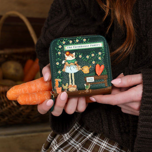 A person in a dark brown knit sweater holds a small embroidered wallet featuring a countryside pantry illustration. The wallet is dark green with floral embroidery, a cartoon girl, and a brown leather base, while the person also holds two carrots in their hands.