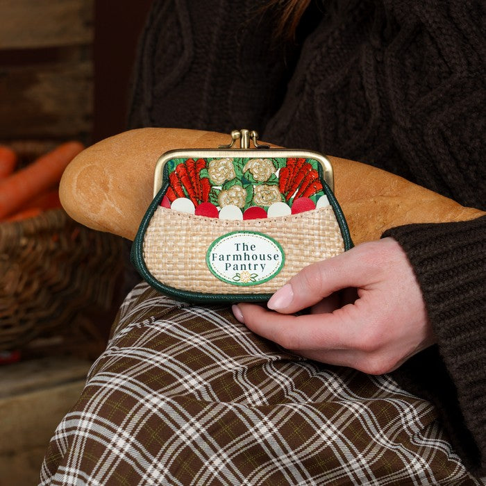 A close-up lifestyle shot of a person holding a small woven coin purse with a vegetable-themed print and a circular 'The Farmhouse Pantry' logo patch. The person is wearing a dark brown cable-knit sweater and a brown and white plaid skirt, with a rustic wooden background.