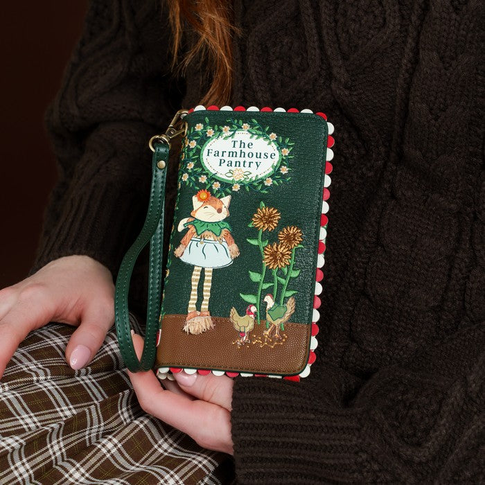 A person in a dark brown cable-knit sweater holds a green faux-leather wallet with a wrist strap. The wallet features a whimsical illustration of a girl in a white dress, sunflowers, chickens, and the text 'The Farmhouse Pantry' inside a white oval.