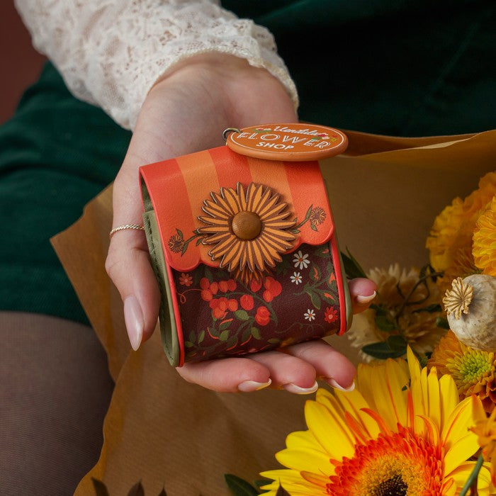 A hand holding a small decorative gift box from 'The Handmade Flower Shop' with a floral pattern and a flower applique, next to a bouquet of yellow flowers.