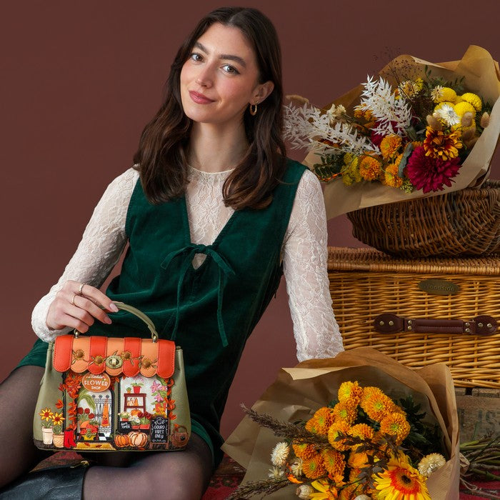 A woman wearing a dark green velvet vest over a white lace long-sleeve top holds a decorative autumn-themed handbag. She is posed in front of a rustic wicker basket filled with dried and fresh flowers against a solid dark brown background.