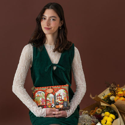 A woman wearing a dark green velvet vest over a white lace long-sleeve top holds a colorful illustrated gift box. She is posed against a solid dark brown background with a bouquet of yellow and orange flowers on the right.