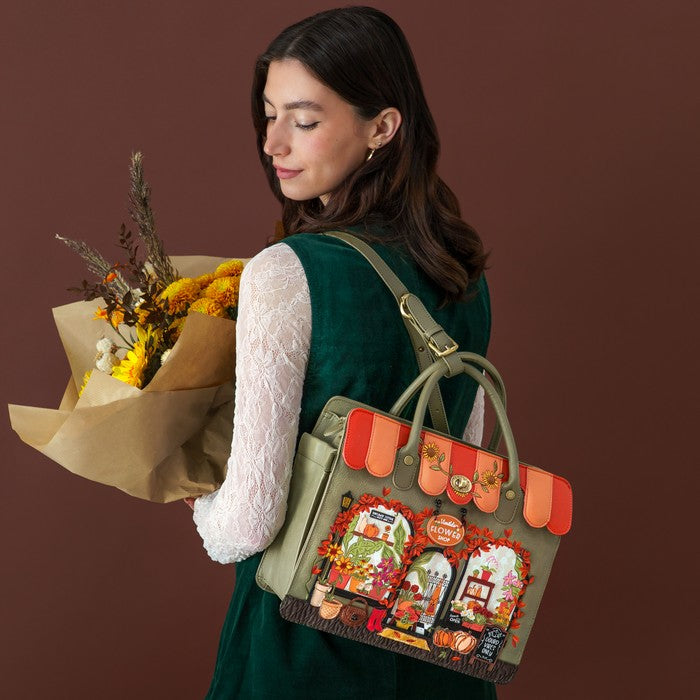 A woman wearing a dark green velvet dress and white lace long-sleeve top carries a large olive green tote bag decorated with autumn-themed illustrations and orange-red striped accents. She is holding a bouquet of yellow and orange flowers against a solid dark brown background.
