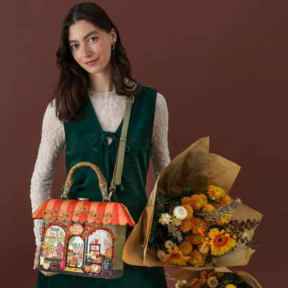 A woman wearing a dark green velvet vest over a cream lace long-sleeve top holds a colorful, whimsical handbag shaped like a street market stall. She is also holding a large bouquet of yellow, orange, and white flowers against a solid dark brown background.