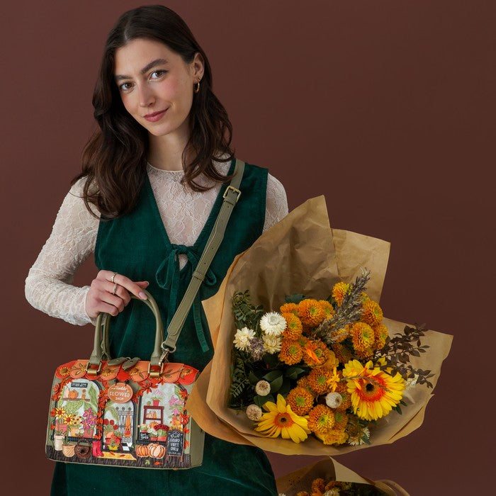 A woman stands against a dark brown studio background holding a colorful illustrated tote bag in one hand and a large bouquet of orange and yellow flowers wrapped in brown paper in the other. She is wearing a white lace long-sleeve top and a dark green pinafore-style dress.