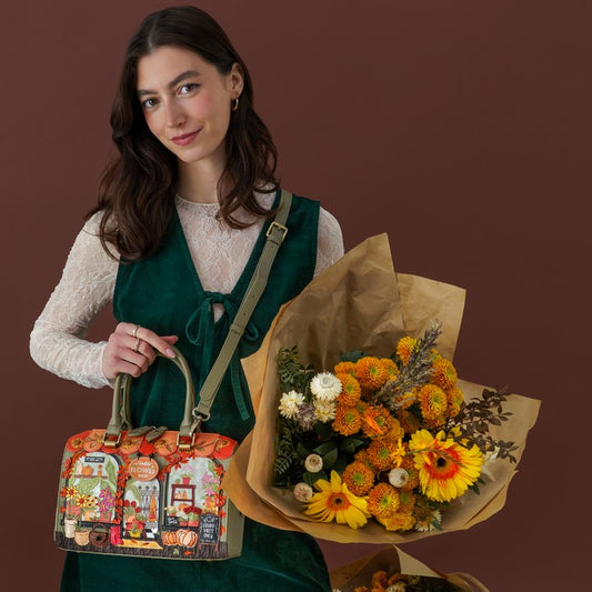 A woman stands against a dark brown studio background holding a colorful illustrated tote bag in one hand and a large bouquet of orange and yellow flowers wrapped in brown paper in the other. She is wearing a white lace long-sleeve top and a dark green pinafore-style dress.
