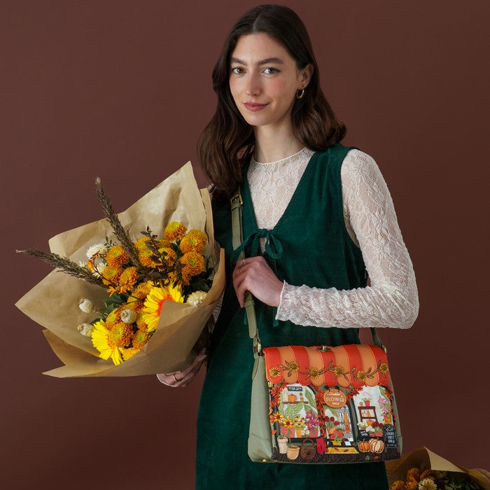 A woman stands against a dark brown studio background holding a bouquet of yellow and orange flowers, wearing a dark green velvet vest over a white lace long-sleeve top and carrying a decorative crossbody bag with a colorful autumn market illustration.