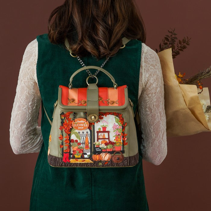 A woman viewed from behind wearing a dark green velvet vest over a white lace long-sleeve top, carrying a small top-handle backpack. The backpack features a colorful embroidered autumn-themed window scene with pumpkins, flowers, and a sign reading 'Happy Halloween'.