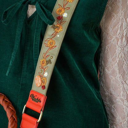 Close-up of a green velvet shoulder strap with colorful floral embroidery, a red leather buckle section, and a visible 'Mukata Designs' logo, worn over a dark green top.