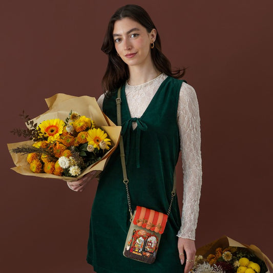 A woman stands against a solid dark brown background wearing a dark green velvet pinafore dress over a white lace long-sleeve top. She is holding a large bouquet of yellow and orange flowers and wearing a small striped crossbody bag with a vintage-style illustration.