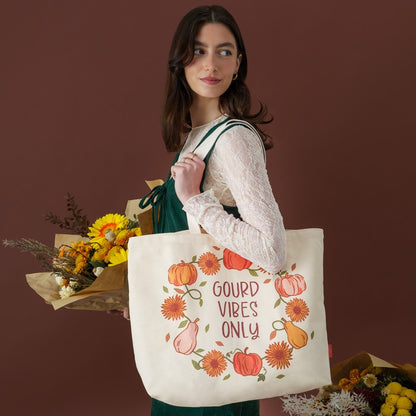 A woman wearing a white lace top and dark green dress carries a cream canvas tote bag with a fall harvest illustration and the text 'GOUD VIBES ONLY'. She is holding dried flowers against a solid dark brown background.