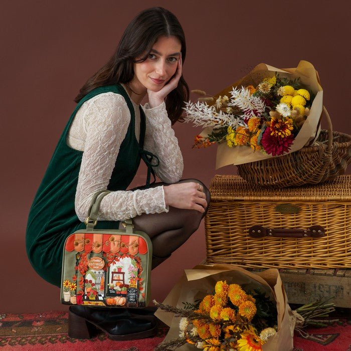A woman crouches beside a vintage wicker trunk and floral arrangements, holding a small illustrated suitcase. She wears a dark green velvet dress over a white lace long-sleeve top.