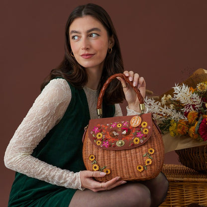A woman in a green velvet vest and white lace top holds a brown woven handbag decorated with colorful floral embroidery and gold-tone hardware, posed against a warm brown studio background with dried flowers and a wicker basket.