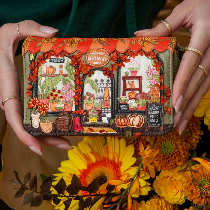 A person holds a small zip-around clutch wallet featuring a highly detailed, colorful autumn-themed illustration of a flower shop. The clutch is decorated with orange leaves, pumpkins, flowers, and a scalloped orange top border.