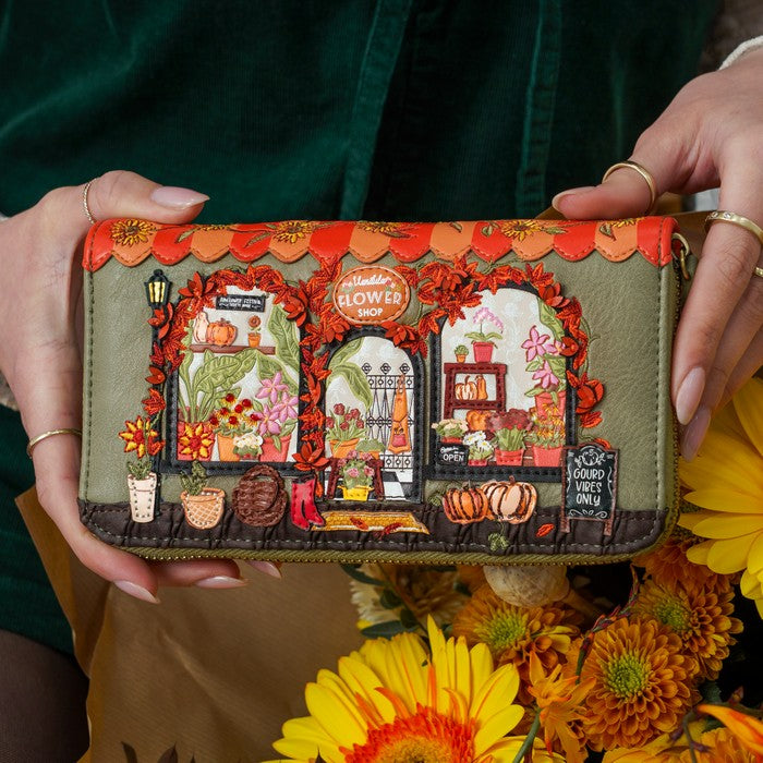 A person holds a small quilted wallet with a detailed autumn flower shop scene. The wallet has olive green base fabric with orange scalloped trim, two arched floral window panels filled with embroidered flowers, pumpkins, and a sign reading 'Wildflower Farm,' and is surrounded by yellow and orange flowers.