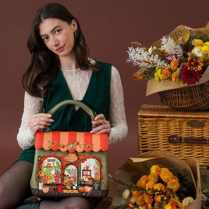 A woman holds a decorative handbag in front of her. The bag has a green base with a colorful illustrated storefront design, orange scalloped trim, and a curved olive green handle.