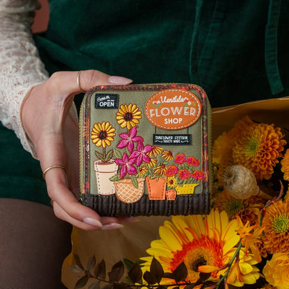A person holds a small zip-around wallet or clutch with floral applique and embroidered details. The front features a patch reading 'Floralista Flower Shop' and illustrations of sunflowers, pink flowers, and potted plants.