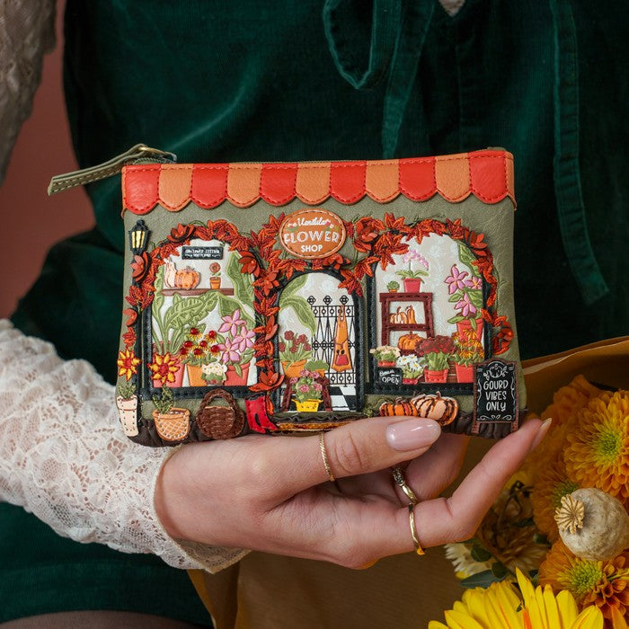 A person holds a small zip pouch featuring an embroidered autumn flower shop scene. The pouch has a beige base with red-orange scalloped trim, orange roof panels, and colorful floral and pumpkin embroidery.
