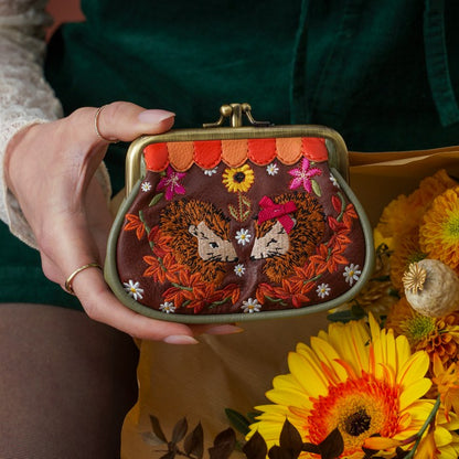 A person holds a small kiss-lock coin purse with intricate floral embroidery. The purse has a dark brown background with embroidered hares, sunflowers, daisies, and orange leaves, along with orange and red scalloped trim.