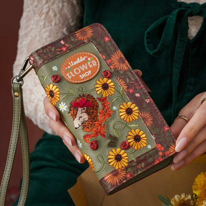 A person holds a decorative zip-around wallet with a wrist strap. The wallet has embroidered floral details, a hedgehog motif, and a round patch that reads 'Ladda's FLOWER SHOP.