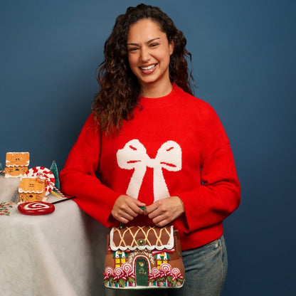 Woman wearing a red sweater with a bow design, standing next to a gingerbread-themed bag and table.