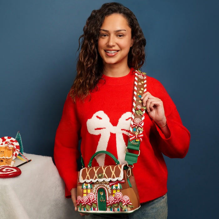 Woman wearing a red sweater with a gingerbread house design, holding a matching handbag against a blue background.