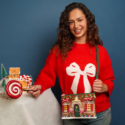 Woman wearing a red sweater with a white bow design, holding a gingerbread house and candy cane against a blue background.