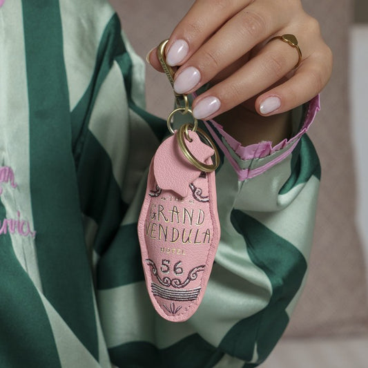 Hand holding a pink keychain with 'Grand Vendula' branding against a green and white striped fabric background.