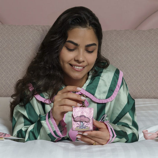 Woman in a green and pink striped robe holding a pink box with a design on it, sitting on a bed.