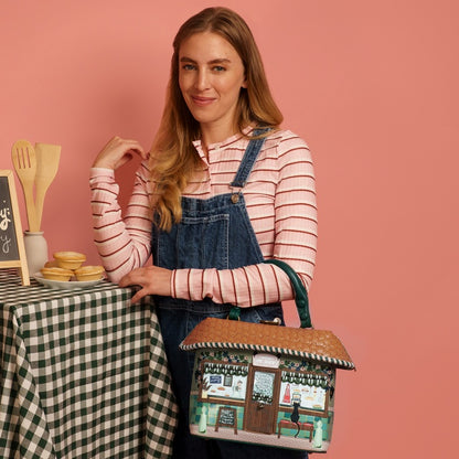 Woman in a striped shirt and denim apron holding a small model of a store against a pink background