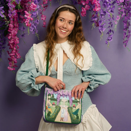 Woman holding a decorative bag with floral and fairy design against a purple background with hanging flowers.