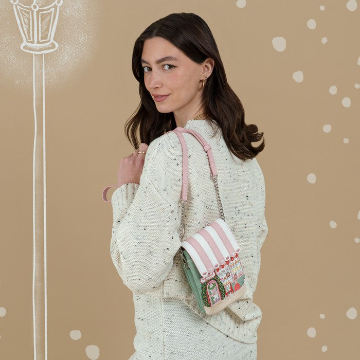 A woman wearing a speckled cream knit sweater carries a small striped crossbody bag with pink and white stripes, a mint green base, and a colorful holiday-themed printed panel on the front, set against a warm beige background with white decorative dots and a faint streetlamp illustration.