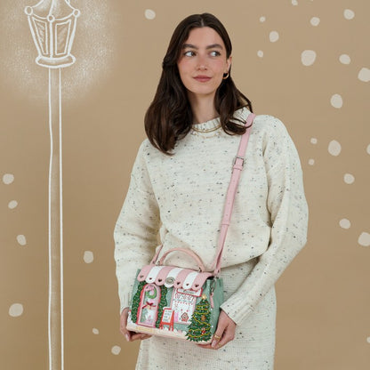 A woman wearing a cream speckled knit sweater and skirt holds a small pink and green holiday-themed handbag with a pink strap against a beige background with white polka dots and a chalk-style street lamp illustration.