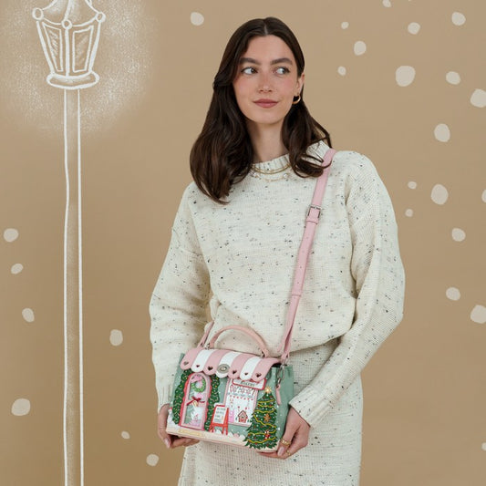 A woman wearing a cream speckled knit sweater and skirt holds a small pink and green holiday-themed handbag with a pink strap against a beige background with white polka dots and a chalk-style street lamp illustration.