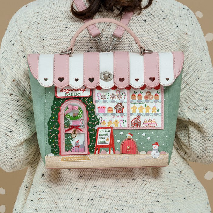 A person wearing a cream knit sweater carries a pastel pink and mint green handbag designed to look like a decorated bakery storefront. The bag has pink and white striped awning details, a green tree-shaped Christmas tree on the left side, and a colorful window display with gingerbread houses, candy canes, and a snowman.
