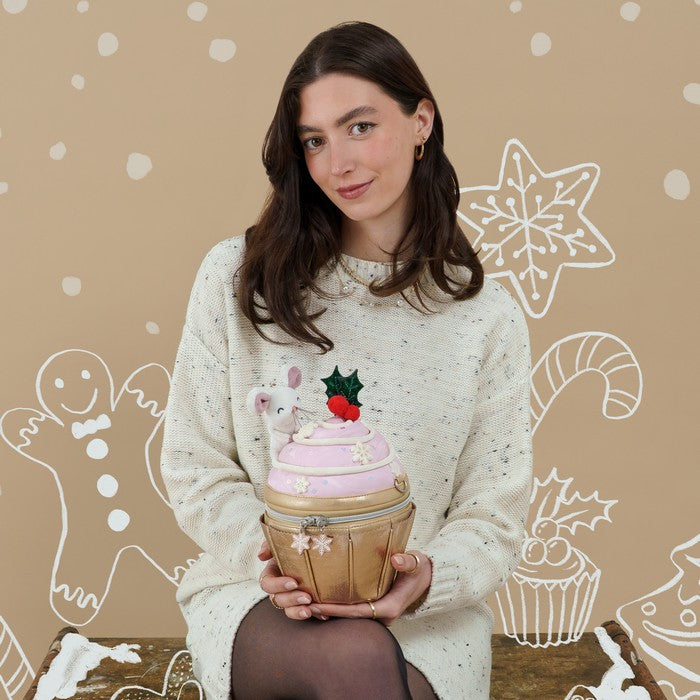 A woman wearing a cream speckled knit sweater holds a cupcake-shaped tin container. The container has pink frosting, a gold cupcake liner, and a small white bunny decoration with a red berry and green holly topper.