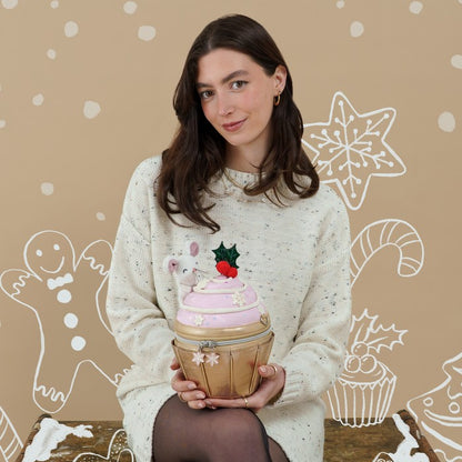 A woman wearing a cream speckled knit sweater holds a cupcake-shaped tin container. The container has pink frosting, a gold cupcake liner, and a small white bunny decoration with a red berry and green holly topper.
