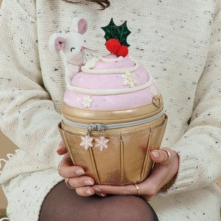 A person in a cream knit sweater holds a cupcake-shaped decorative box. The box has a pink frosting top with a white mouse figurine, red berries, and green holly, set in a gold wrapper-like base with small white flower details.