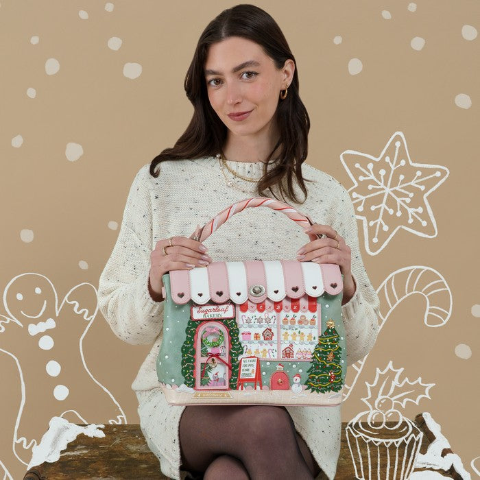 A woman wearing a cream knit sweater holds a decorative Christmas-themed handbag shaped like a candy store. The bag has a pink and white striped awning, a green body, and festive details including a Christmas tree, candy cane, and gift boxes.