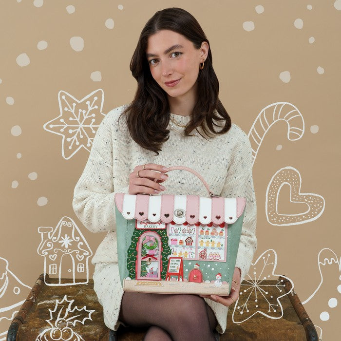 A woman wearing a cream knit sweater holds a decorative handbag styled like a candy store. The bag has a pink and white striped awning, a mint green body, and various candy-themed illustrations, set against a beige background with white holiday doodles.